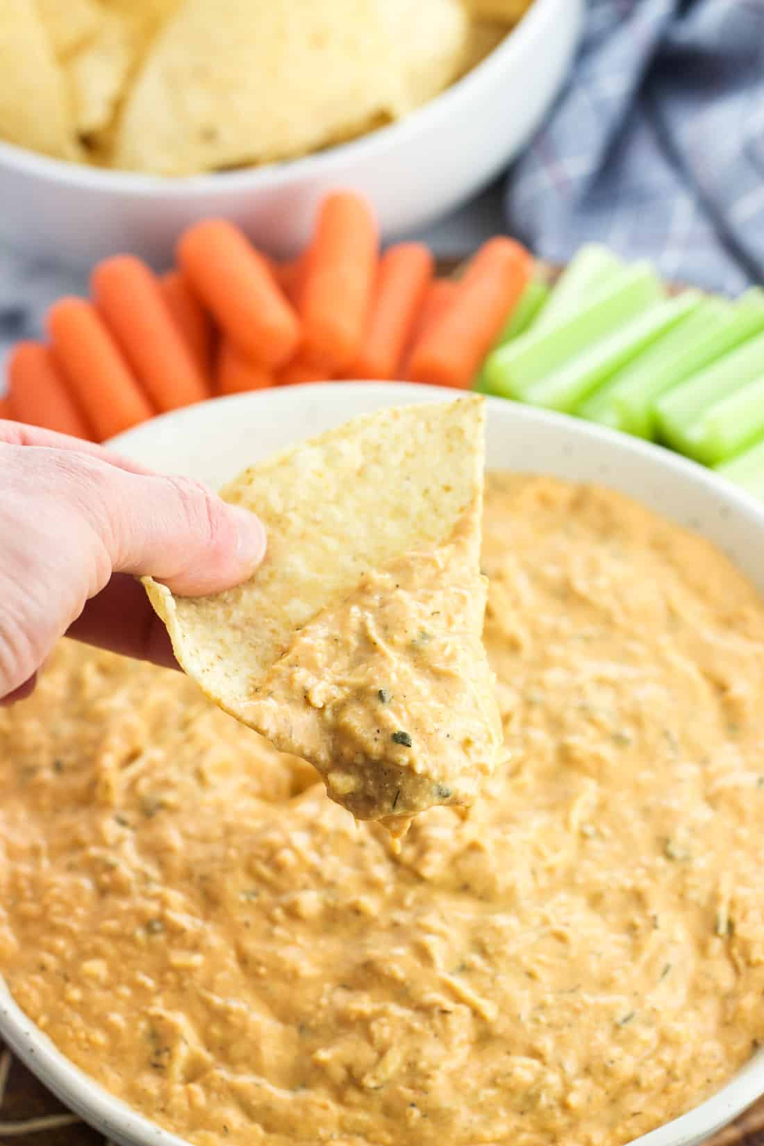 A hand lifting a tortilla chip out of the serving bowl after scooping dip.