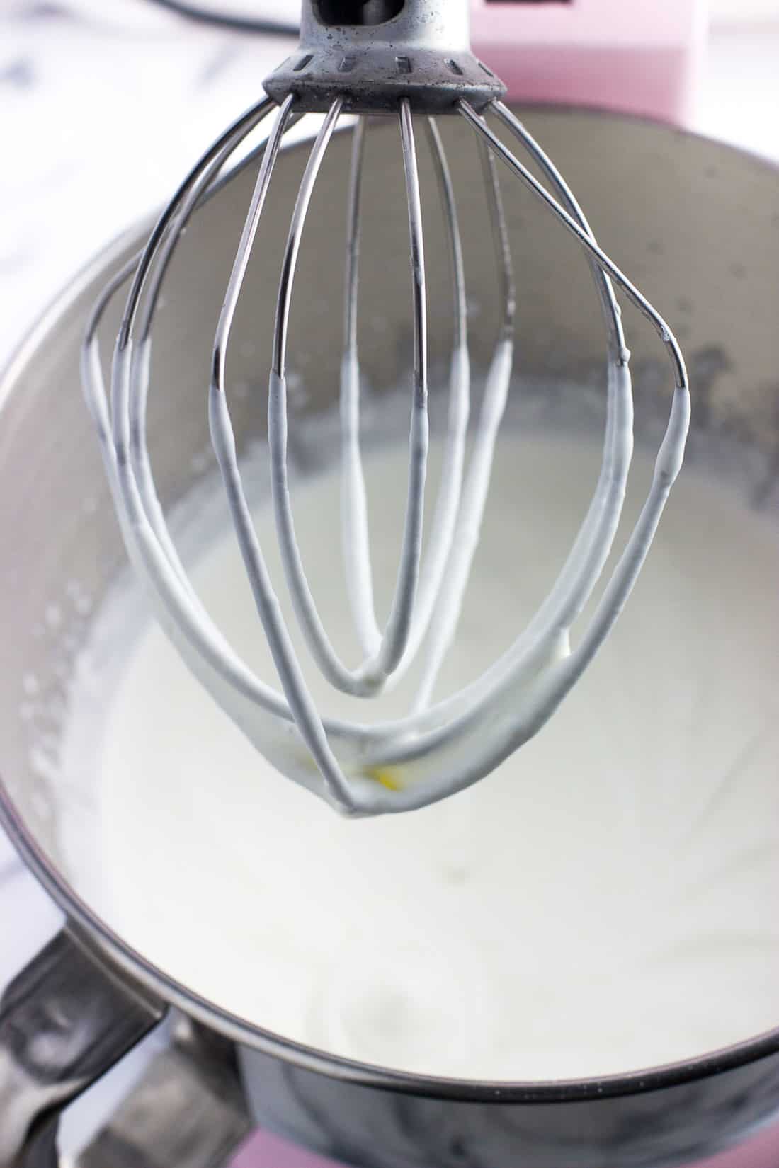 A stand mixer whisk attachment lifted out of the whipped cream mixture as it begins to thicken.