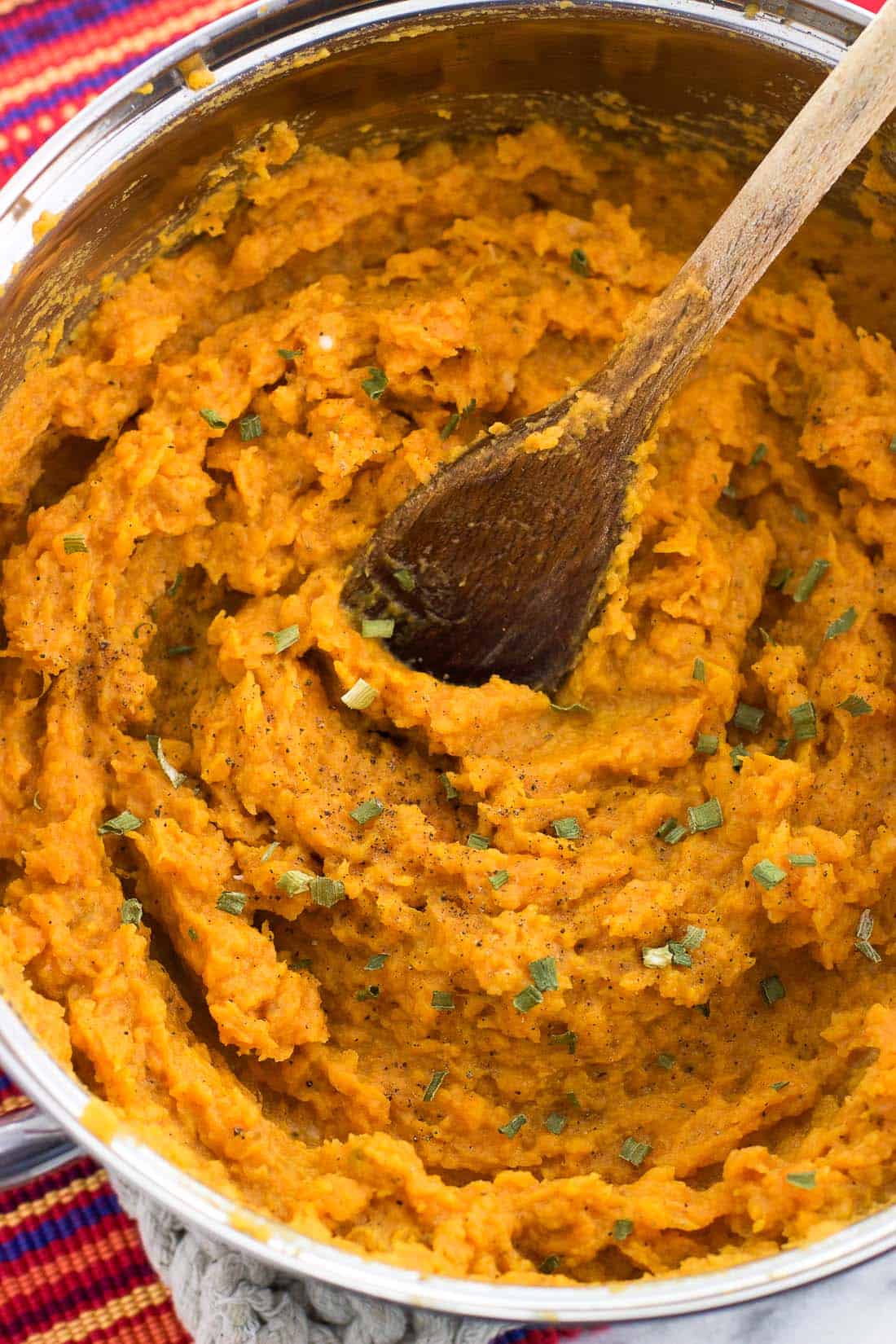 Mashed sweet potatoes in a pan with a wooden spoon and garnished with chives