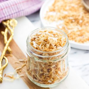 A small glass jar of toasted coconut on a marble board in front of a big plate of more coconut flakes