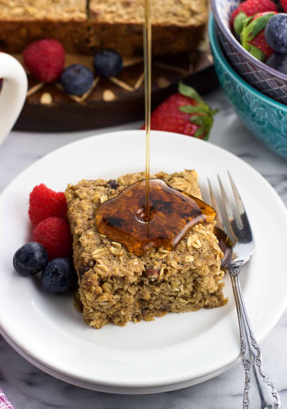 Fresh fruit and a baked oatmeal square on a plate being drizzled by maple syrup.