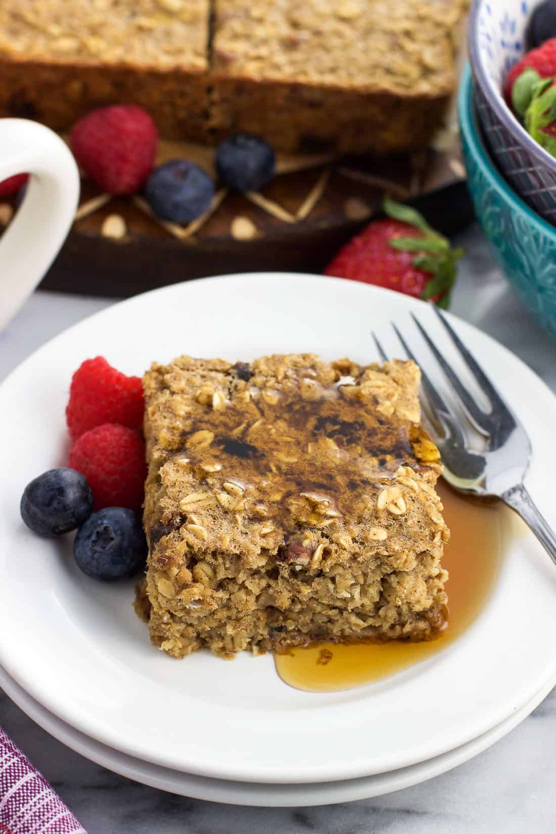 A maple syrup drizzled square of baked oatmeal on a plate with a fork and berries.