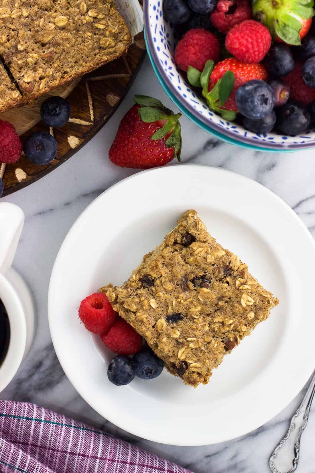 A piece of baked oatmeal on a plate next to a cup of coffee and a bowl of fruit.