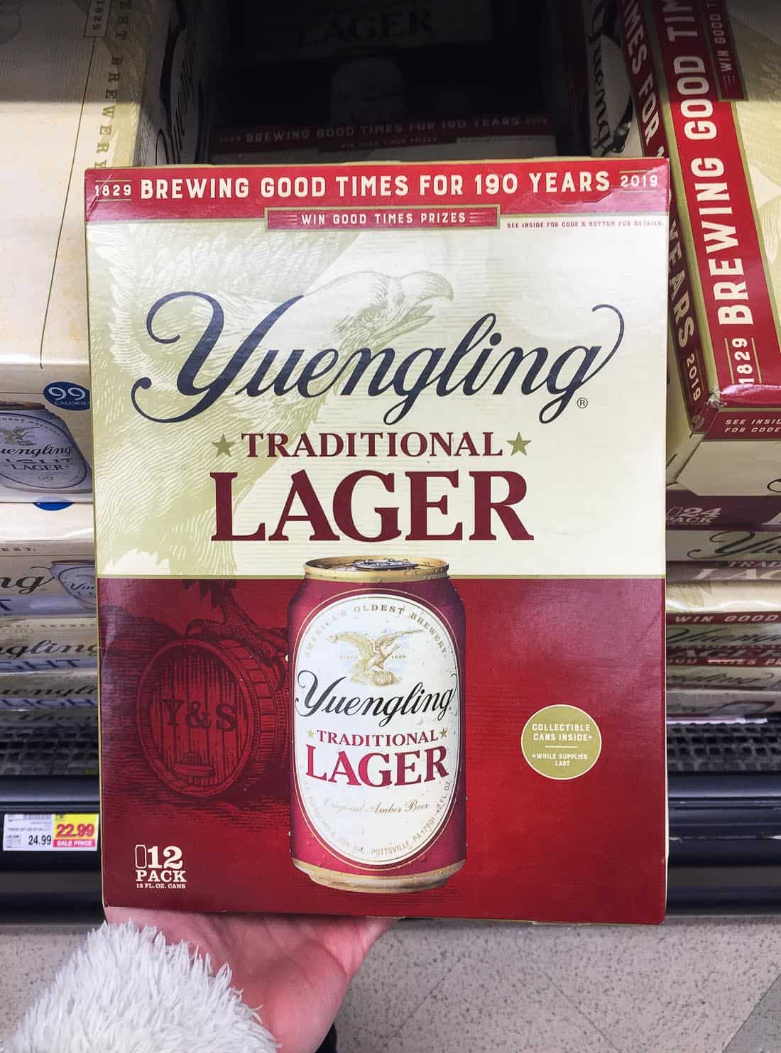 A hand holding a 12-pack of Yuengling Lager in the grocery store beer section.