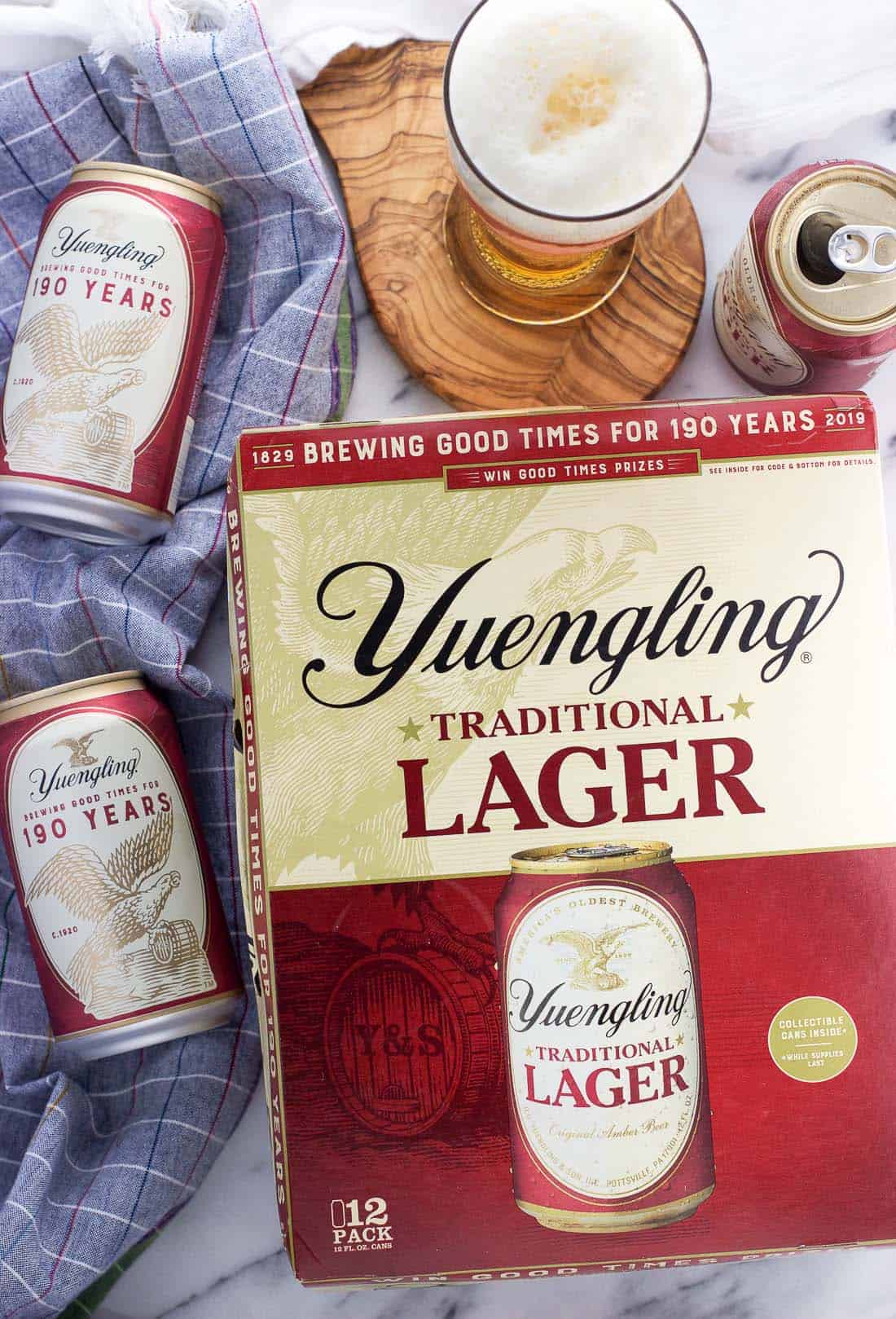 An overhead picture of a 12-pack of Yuengling Lager surrounded by cans of beer and a foamy-headed glass of beer.