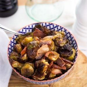 A small ceramic bowl on a wooden serving board filled with roasted brussels sprouts and bacon