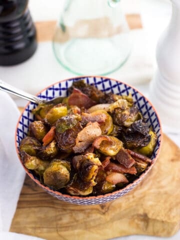 A small ceramic bowl on a wooden serving board filled with roasted brussels sprouts and bacon