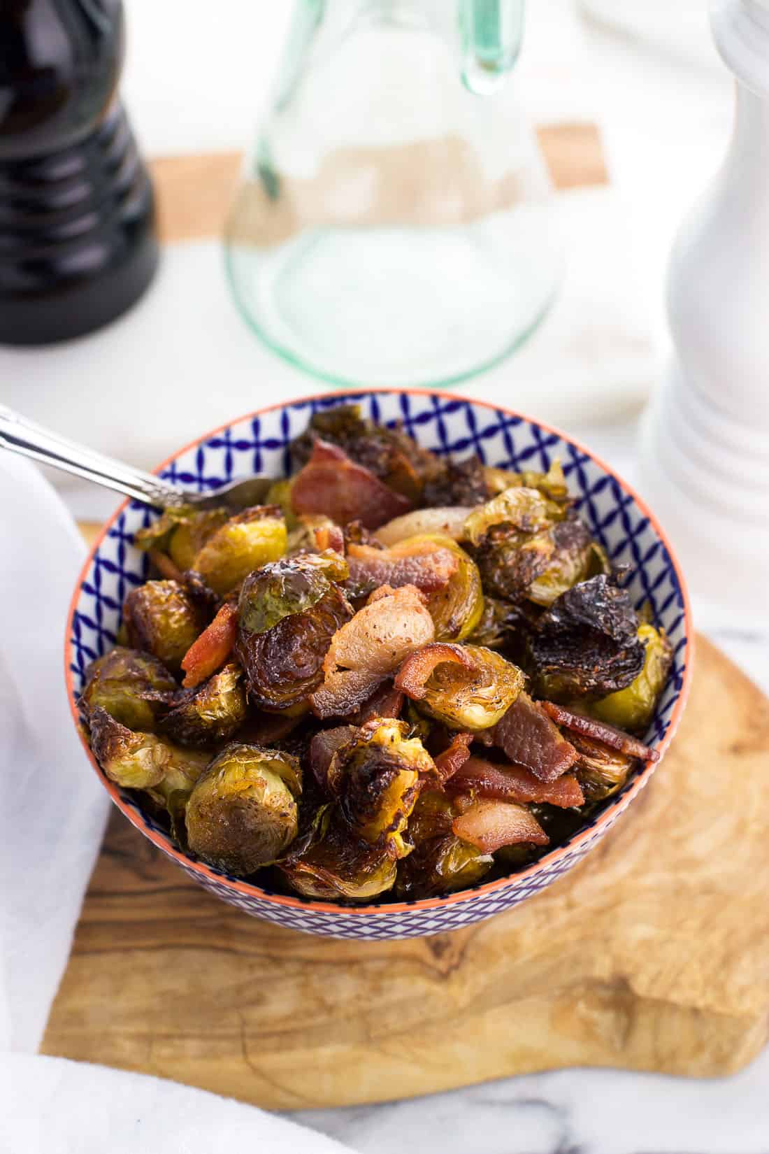 A small ceramic bowl on a wooden serving board filled with roasted brussels sprouts and bacon