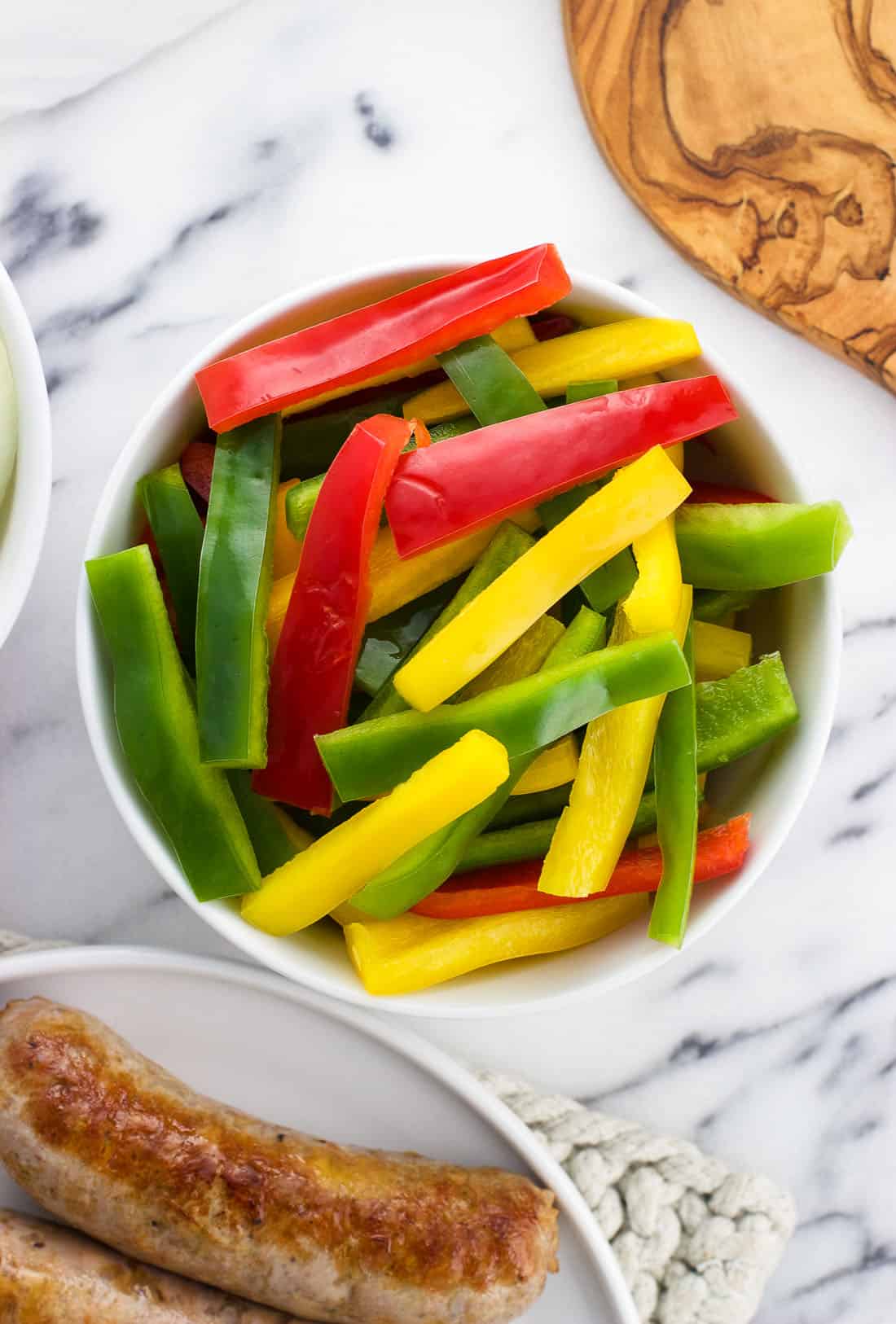 A variety of raw sliced peppers in a bowl next to browned Italian sausages.