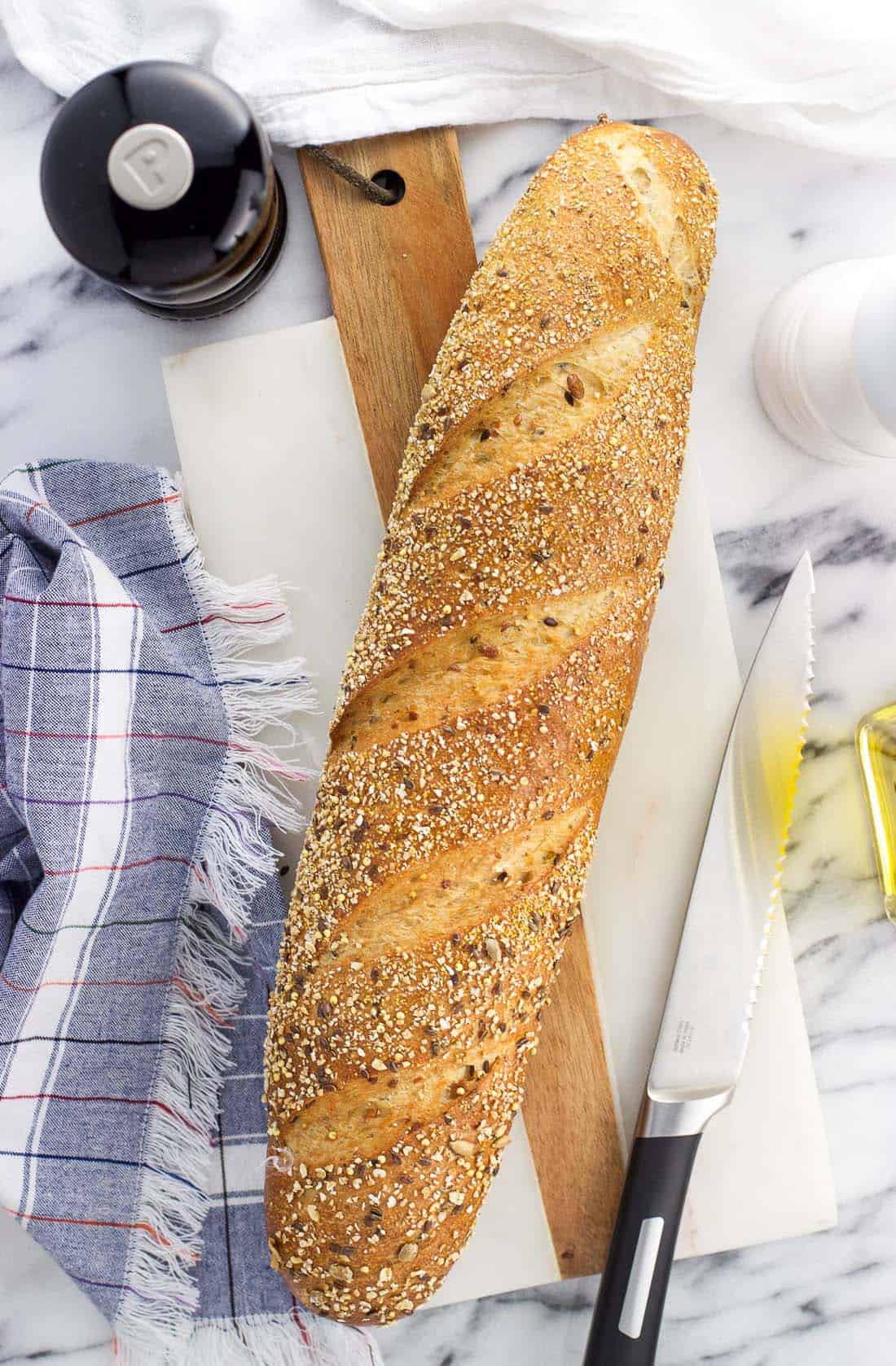 An overhead shot of a baguette loaf, salt and pepper grinders, and olive oil on a marble board.