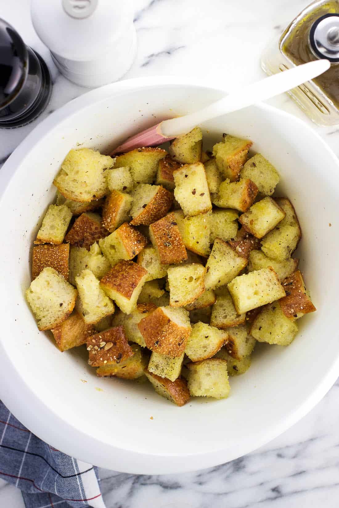 Bread cubes tossed with olive oil, salt, and pepper with a spatula in a mixing bowl.