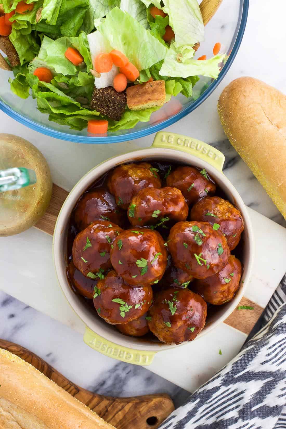 An overhead shot of glazed BBQ chicken meatballs in a bowl next to a bowl of salad and bread rolls.