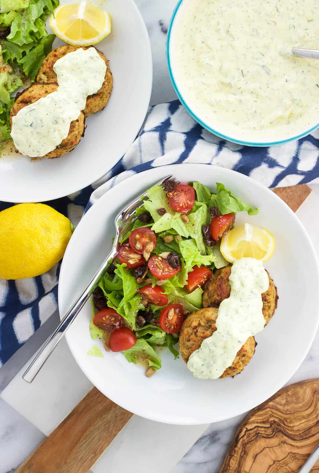 An overhead shot of a plate of two tuna cakes, a drizzle of tartar sauce, a lemon wedge, and a side salad.