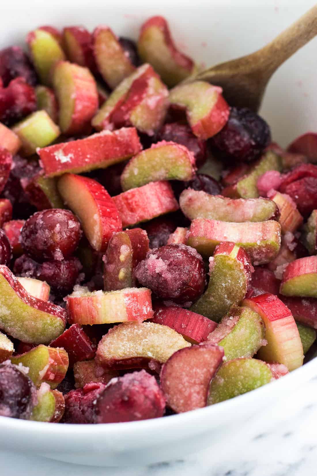 Sliced rhubarb and cherries covered in a sugar mixture in a mixing bowl