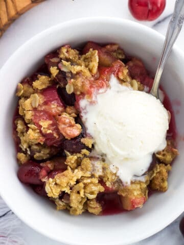 An overhead shot of a serving of cherry rhubarb crisp in a bowl with a scoop of vanilla ice cream and a spoon