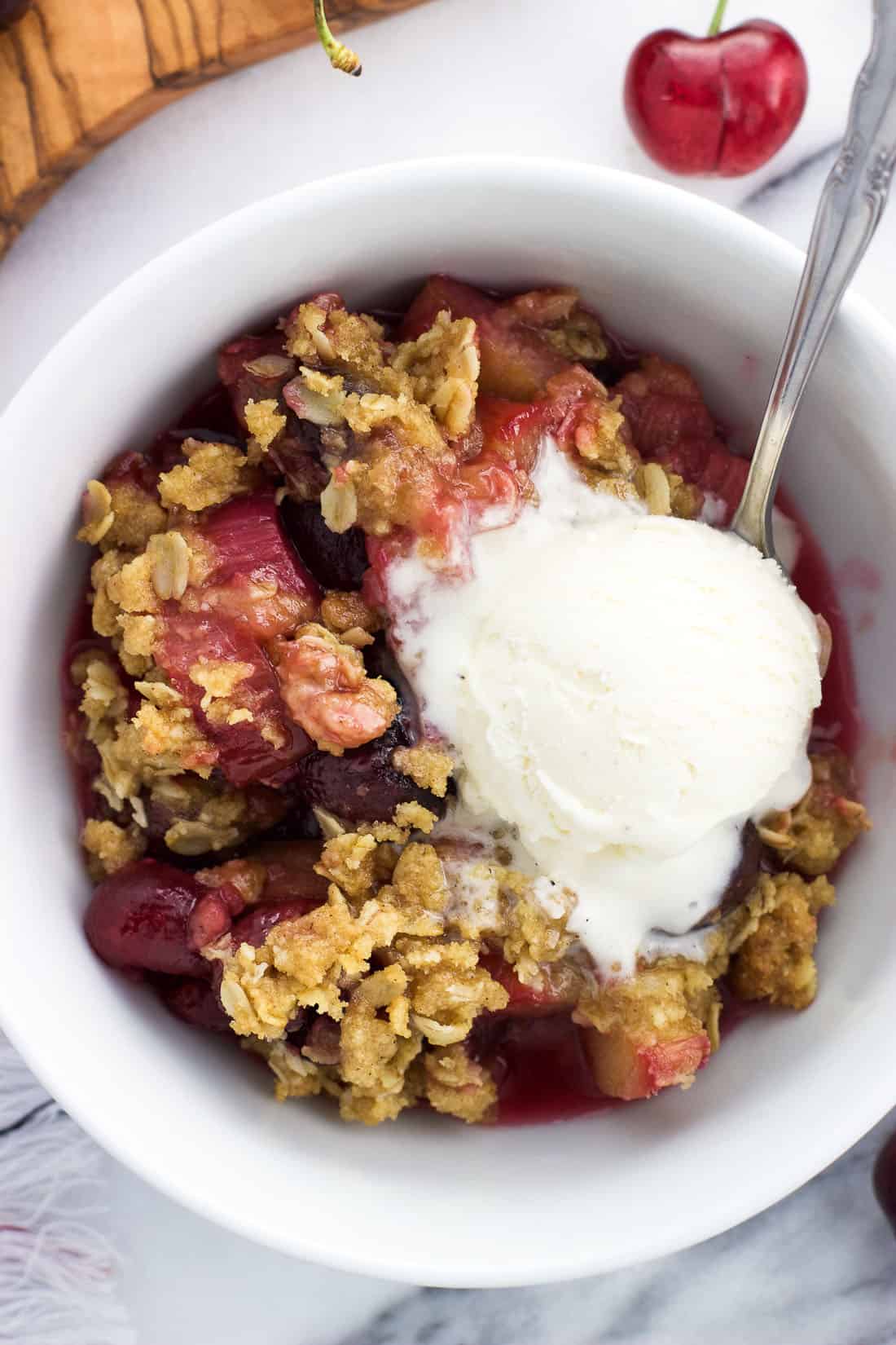 An overhead shot of a serving of cherry rhubarb crisp in a bowl with a scoop of vanilla ice cream and a spoon