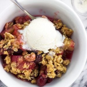 An overhead shot of a serving of cherry rhubarb crisp in a bowl with a spoon and a scoop of vanilla ice cream