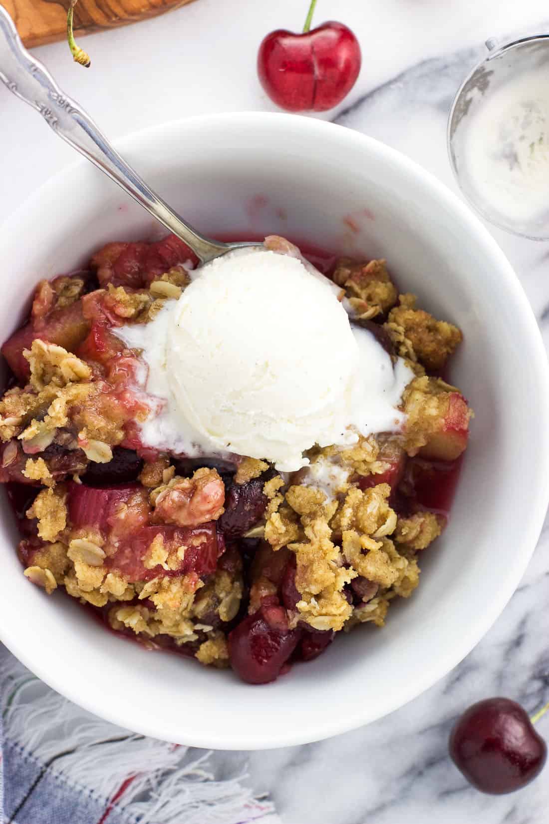 An overhead shot of a serving of fruit crisp in a bowl with a spoon and a scoop of vanilla ice cream