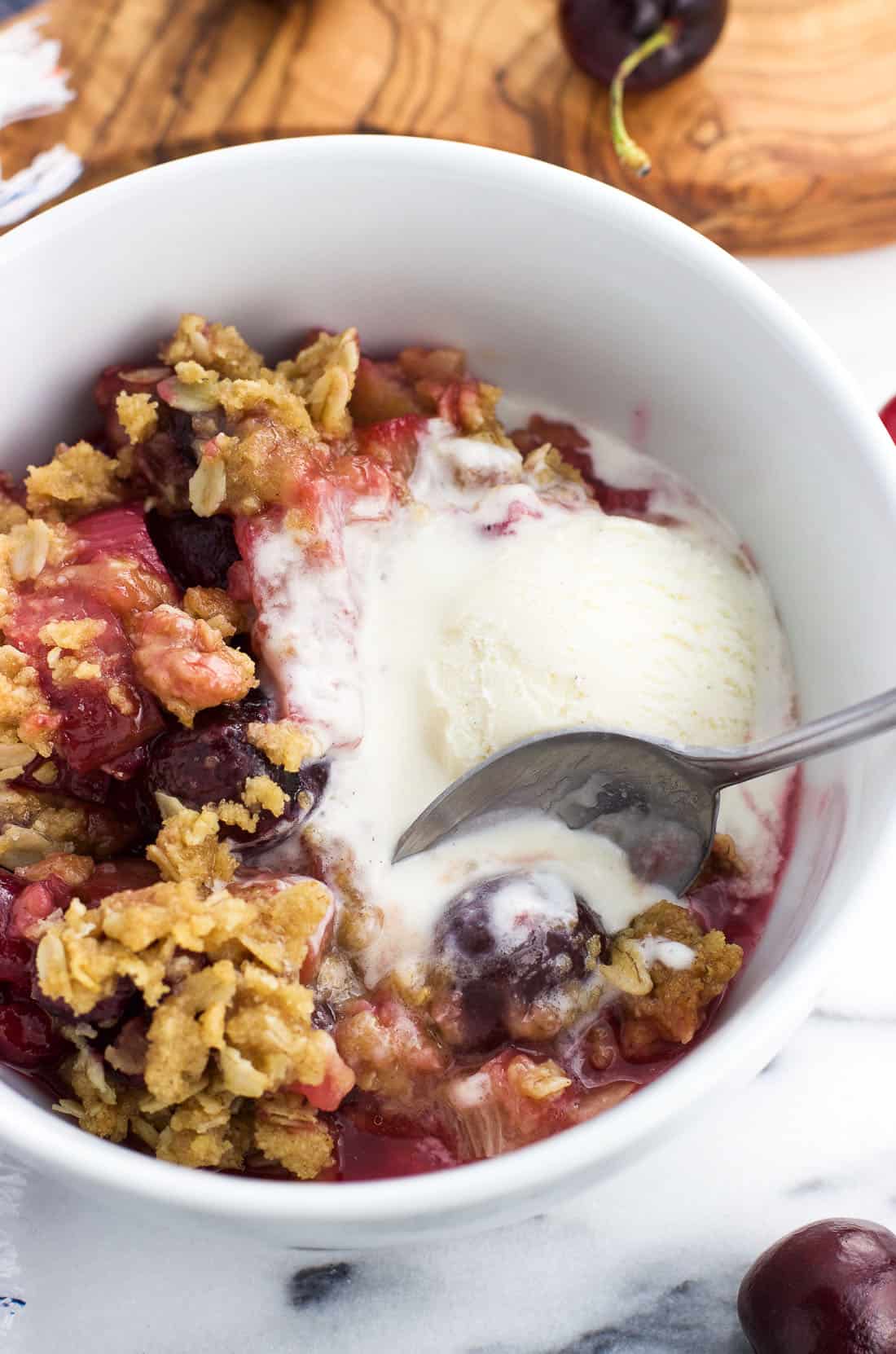 A spoon scooping up a cherry with vanilla ice cream from a serving of fruit crisp in a bowl