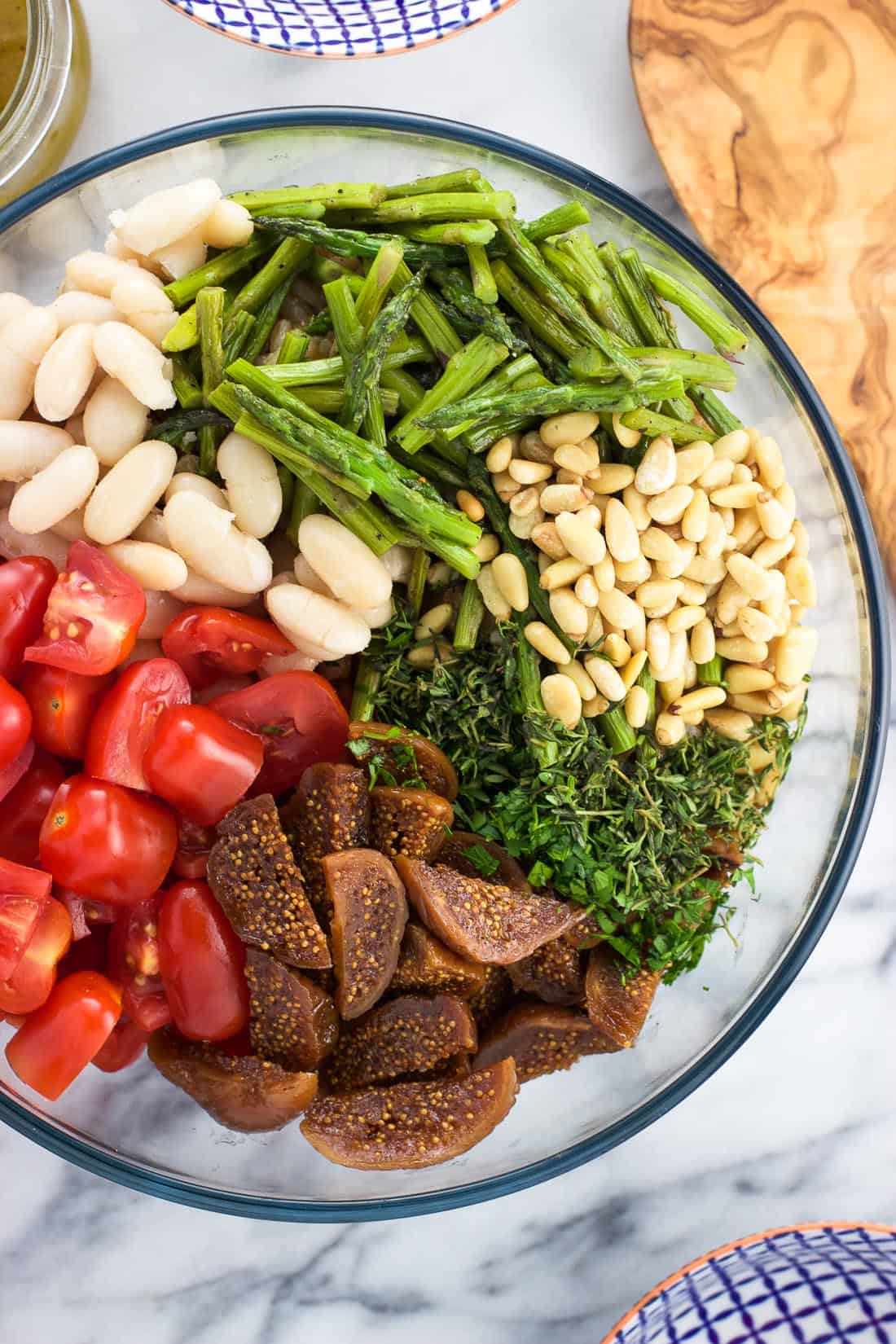 An overhead shot of tomatoes, white beans, asparagus, pine nuts, herbs, and figs on top of farro in a serving bowl before being mixed together.