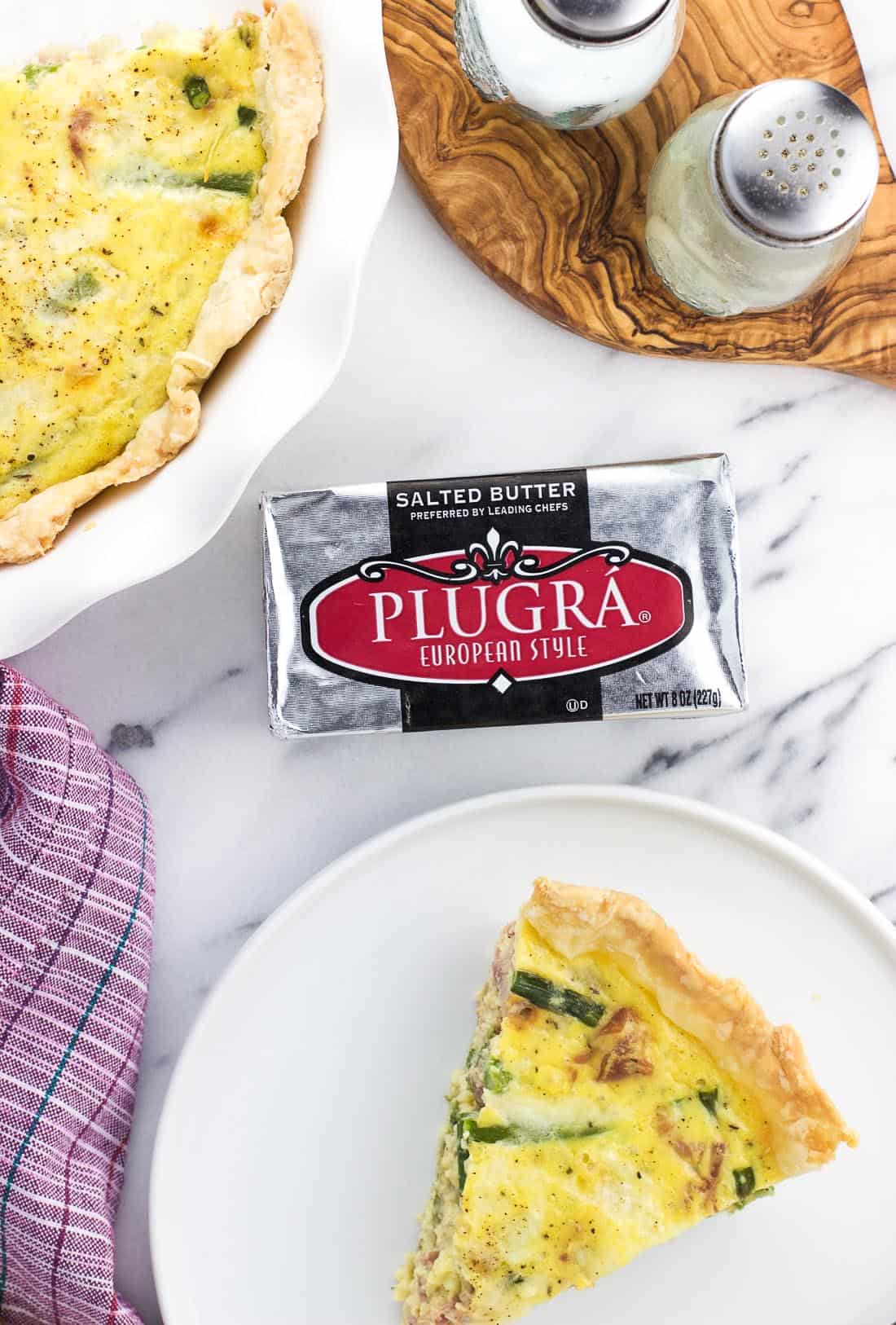 A package of Plugra butter on a marble board surrounded by a slice of quiche on a plate and the rest of the quiche in a pie plate.