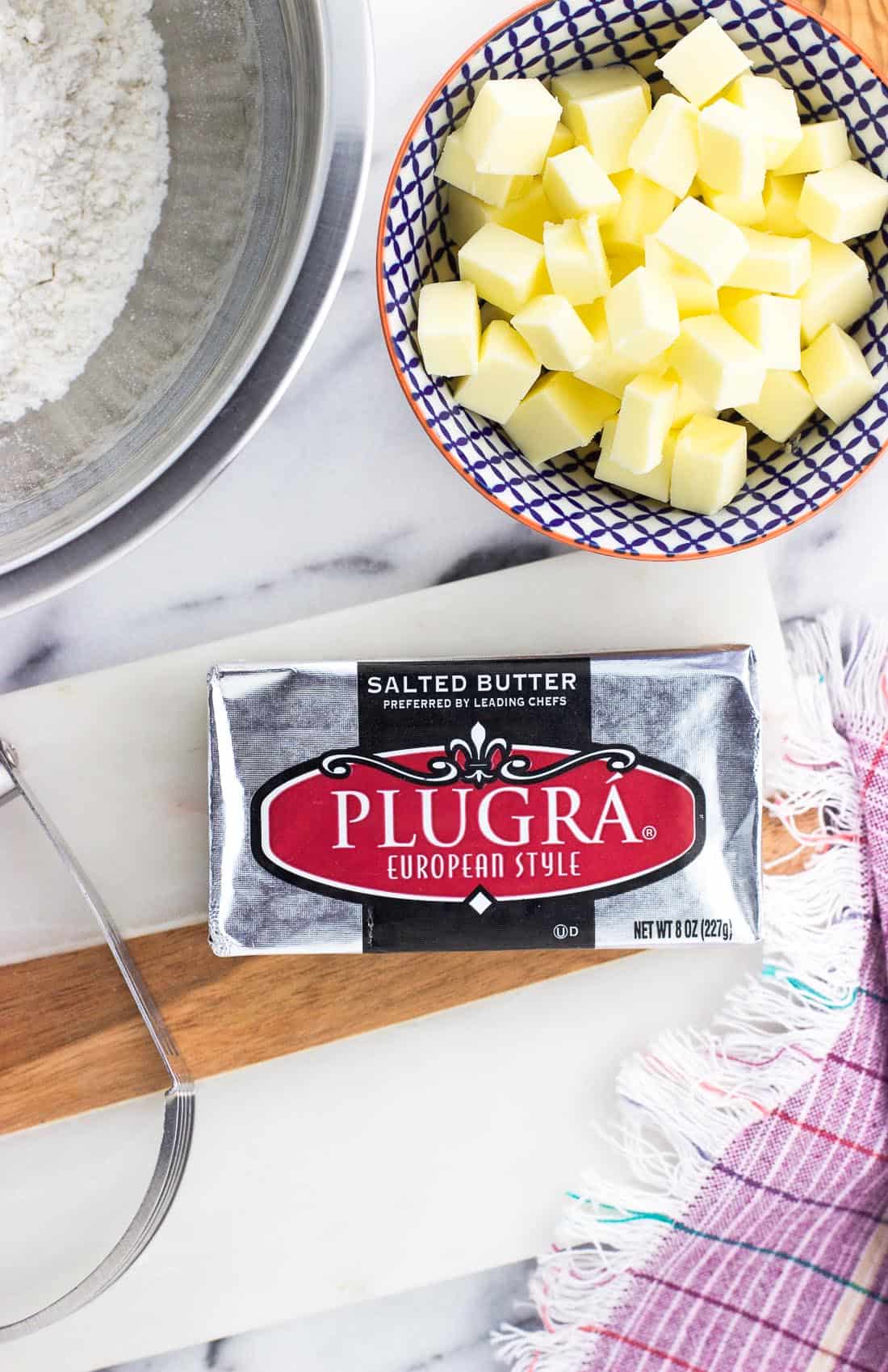 A package of Plugra butter on a marble board surrounded by a bowl of flour, a pastry blender, and a bowl of diced butter.
