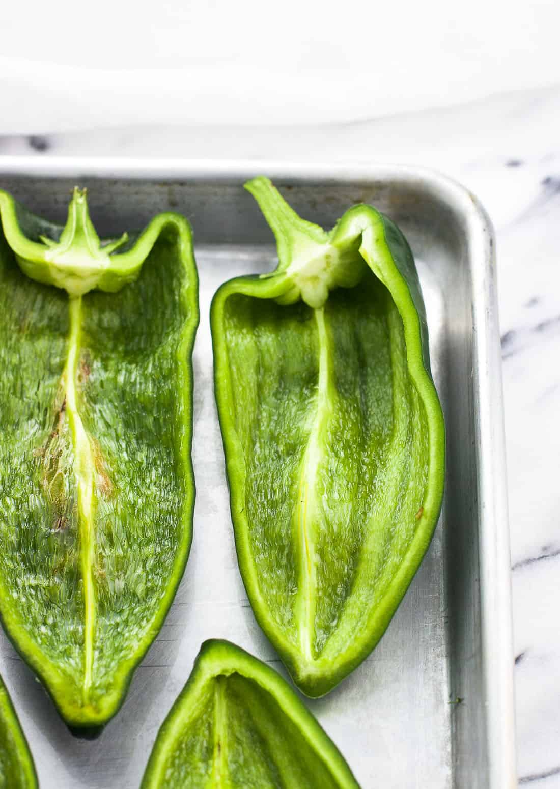 Raw poblano peppers halved and lined up next to one another on a metal baking sheet.