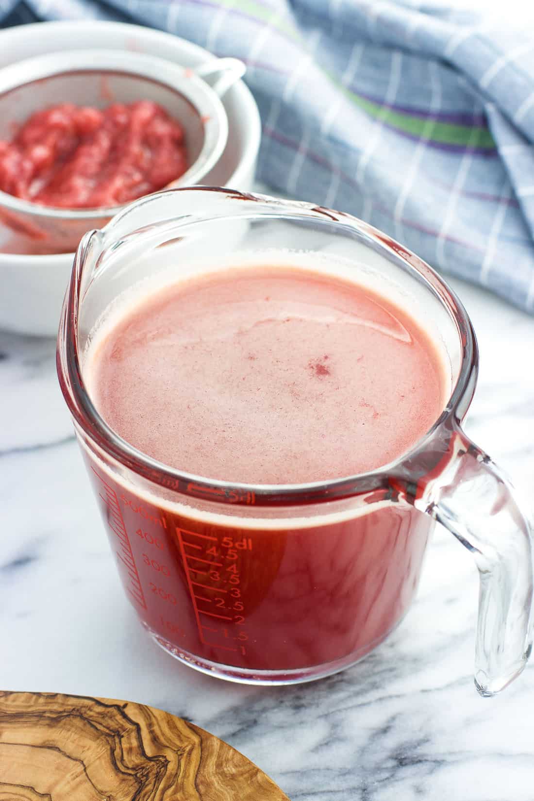 Strawberry syrup in a glass measuring cup with a strainer of mashed strawberries over a bowl in the background