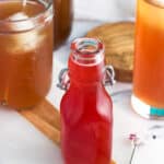 A small glass jar of strawberry syrup with glasses of iced tea in the background
