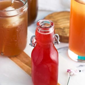 A small glass jar of strawberry syrup with glasses of iced tea in the background