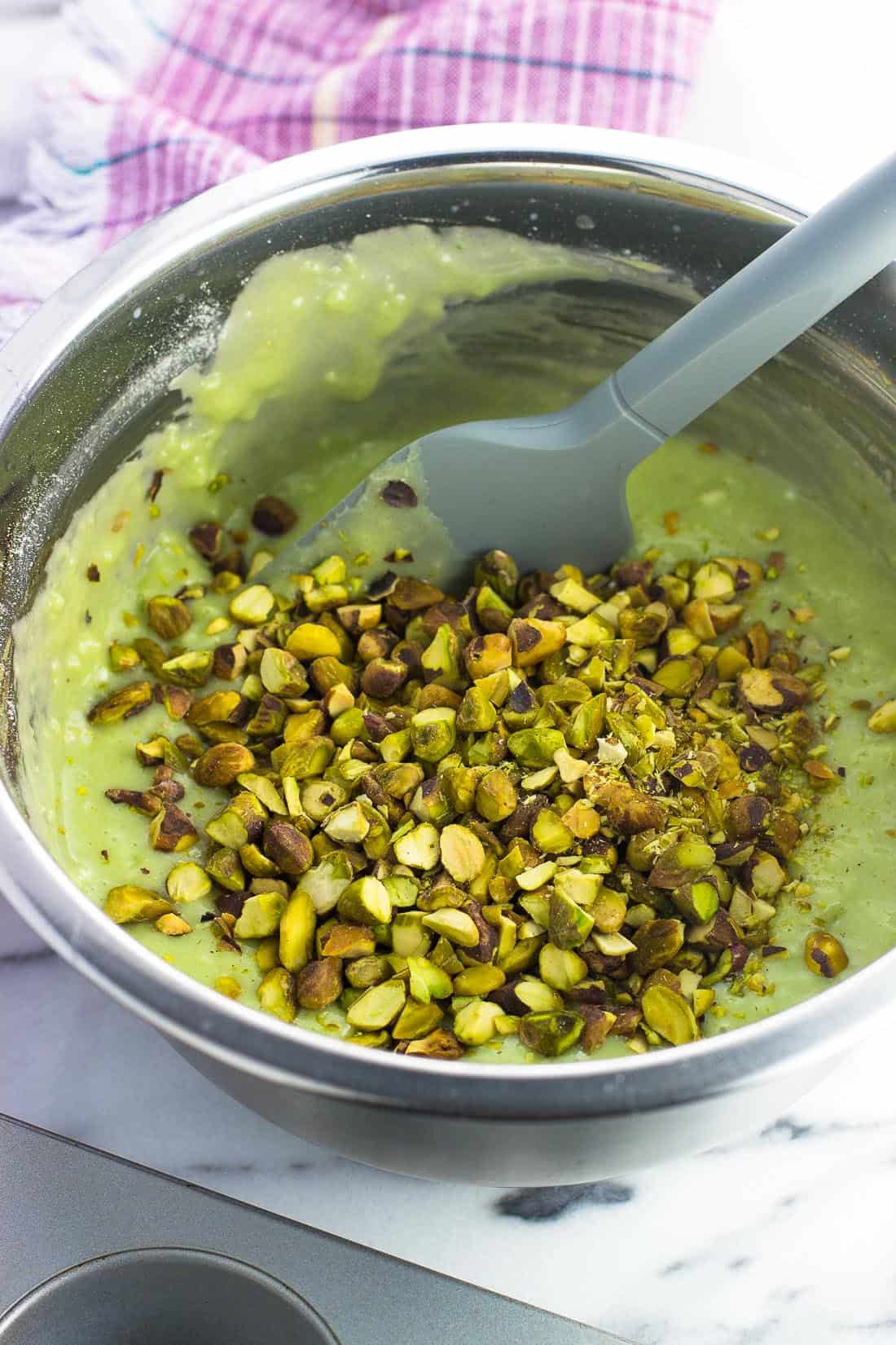 Pistachio muffin batter in a metal mixing bowl with chopped pistachios ready to be stirred in