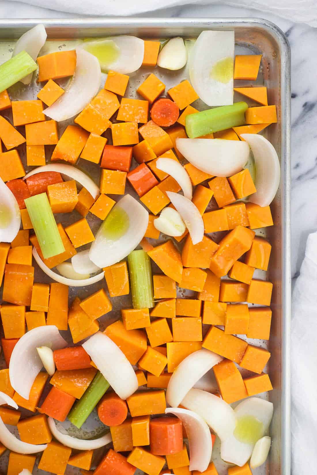 Butternut squash soup ingredients on a baking sheet before roasting, including butternut squash cubes, carrots, celery, onion, and garlic cloves