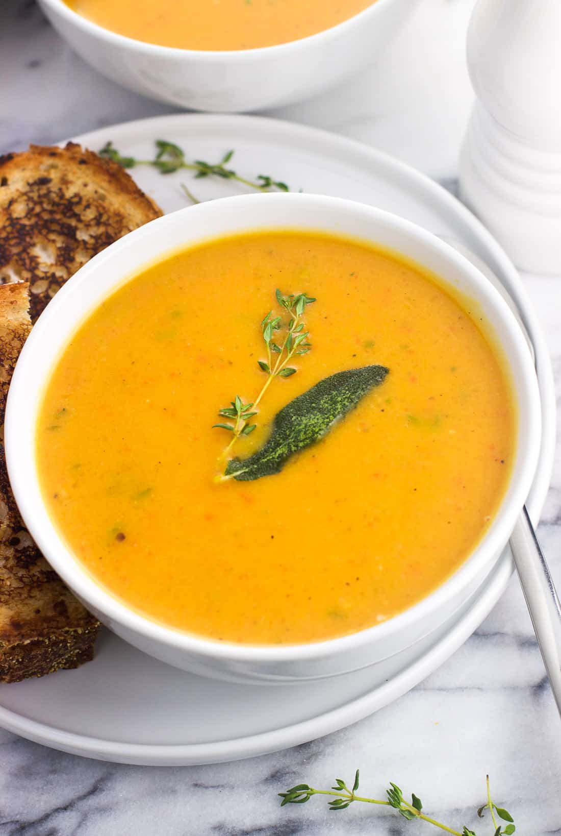 A bowl of roasted butternut squash soup garnished with a sprig of thyme and a sage leaf, served next to a grilled cheese sandwich.