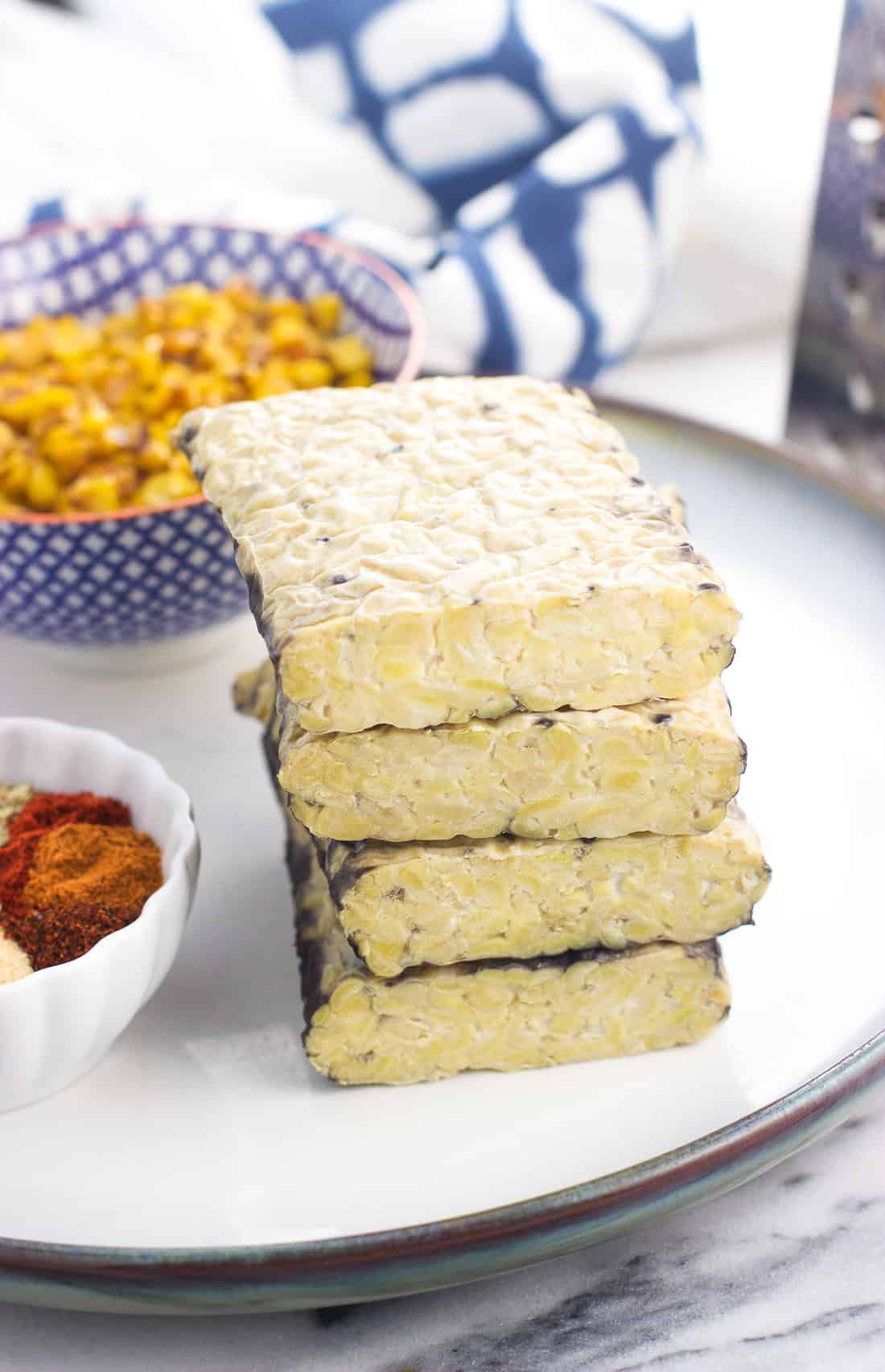 Blocks of tempeh stacked on top of one another on a ceramic plate next to taco seasoning and roasted corn