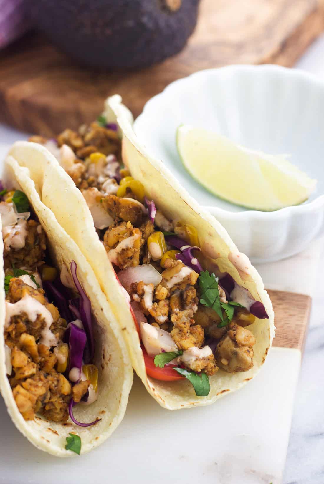 Tacos filled with tempeh, onion, tomatoes, cabbage, corn, cilantro, and more standing next to one another on a serving board