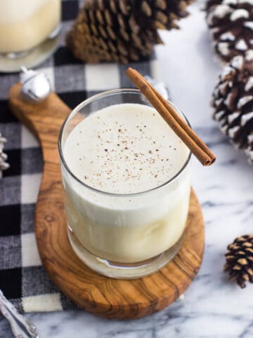 A glass of cooked eggnog on a wooden board garnished with a cinnamon stick