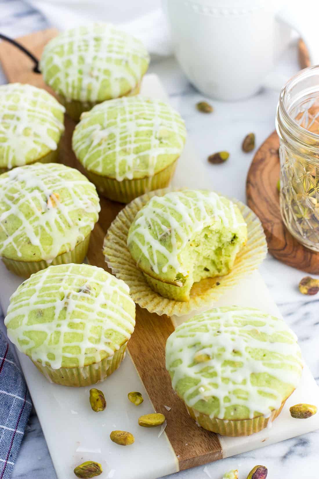 Pistachio muffins on a marble serving board. One muffin has the paper peeled back with a bite taken out of it