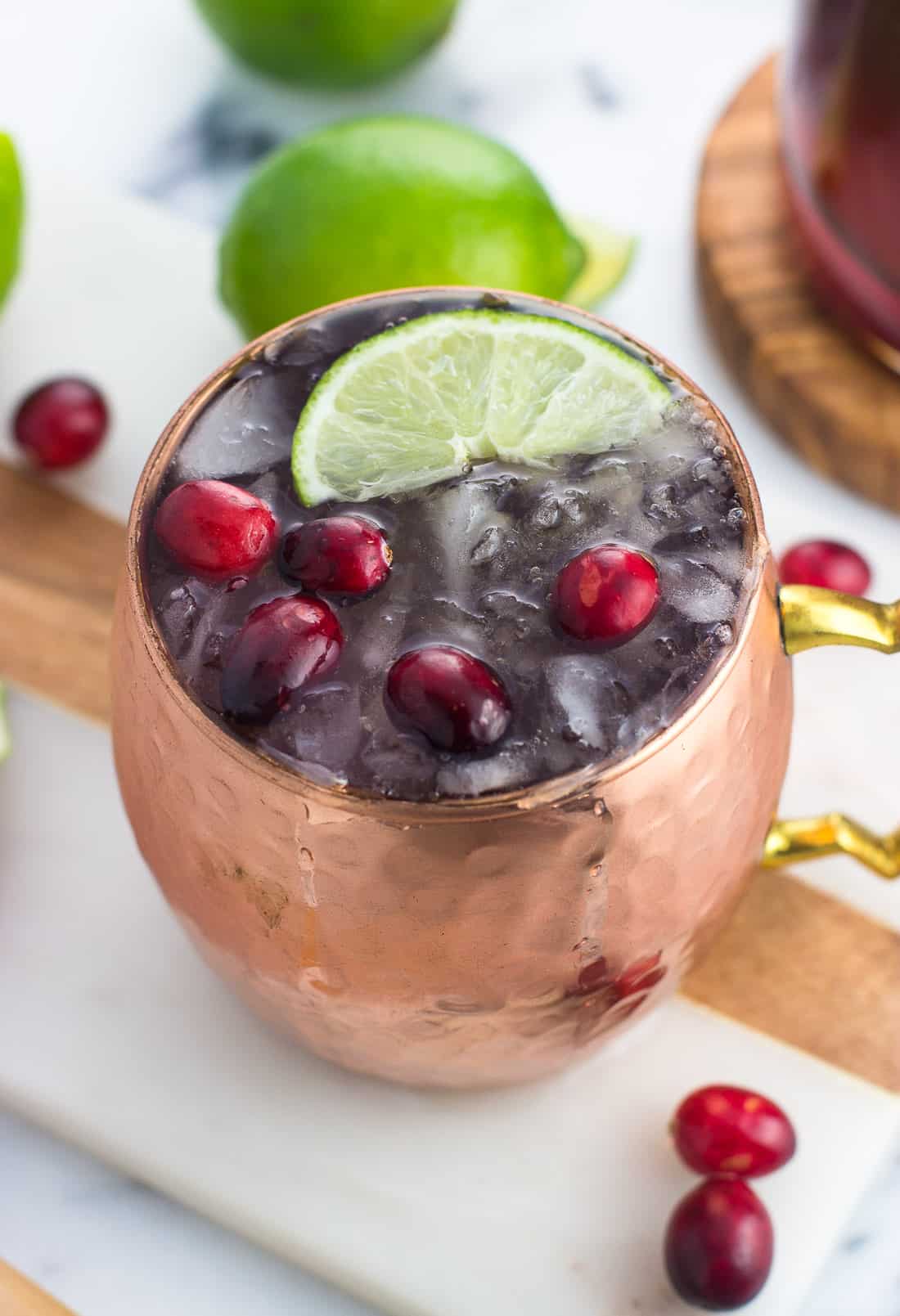 A cocktail in a mug on a marble serving board surrounded by fresh cranberries and limes