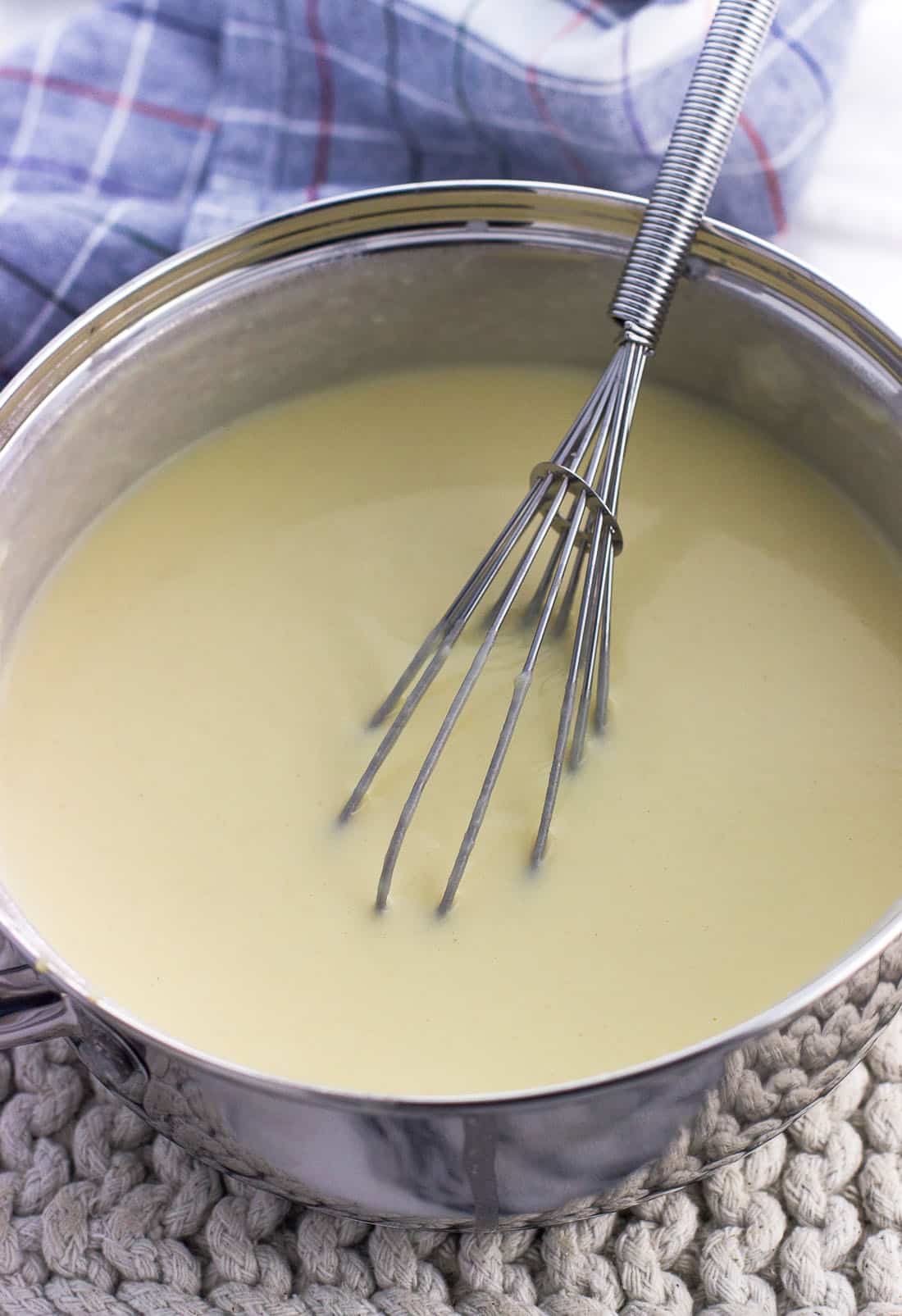 The roux in a small saucepan with milk whisked in and a metal whisk.