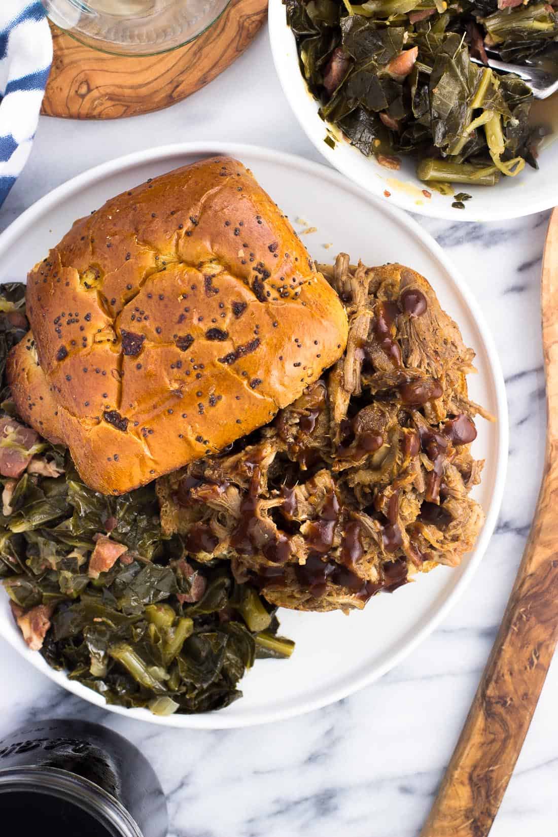 An overhead picture of a pulled pork sandwich on an onion roll on a plate with a side of southern greens