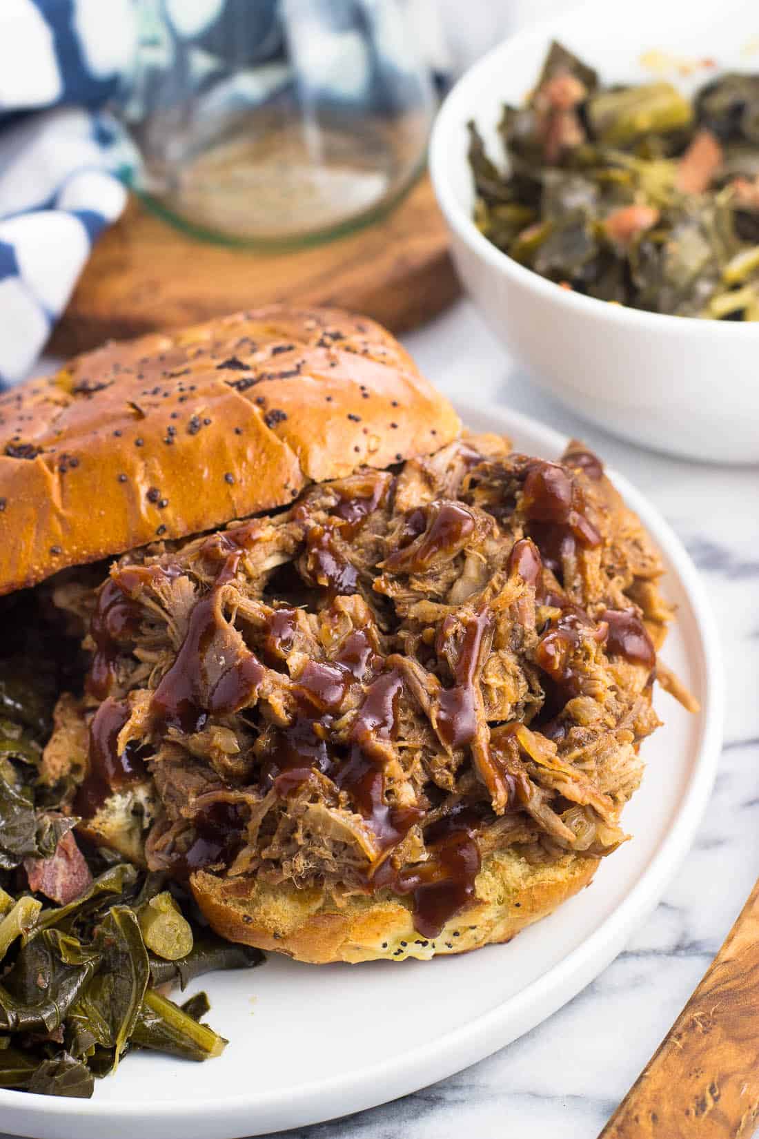 Slow cooker pulled pork drizzled with BBQ sauce on an onion roll served next to southern greens