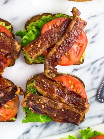 Four pieces of avocado toast on a marble board, topped with lettuce, tomato slices, and candied bacon