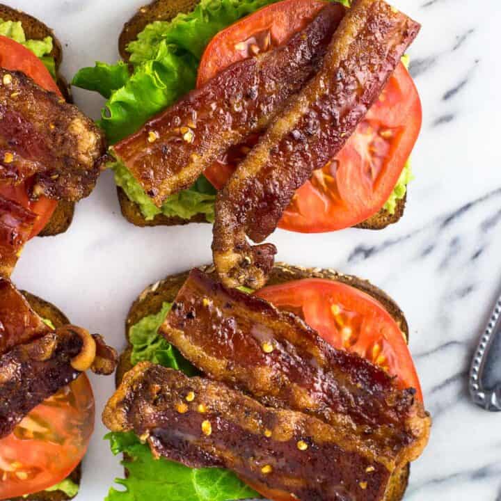Four pieces of avocado toast on a marble board, topped with lettuce, tomato slices, and candied bacon