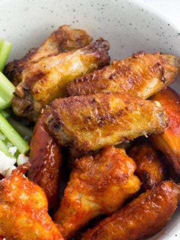 A bowl filled with chicken wings (half plain and half coated in hot sauce) with celery sticks, baby carrots, and crumbled blue cheese