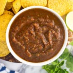 An overhead picture of a bowl of salsa on a wooden board surrounded by tortilla chips and a bunch of cilantro