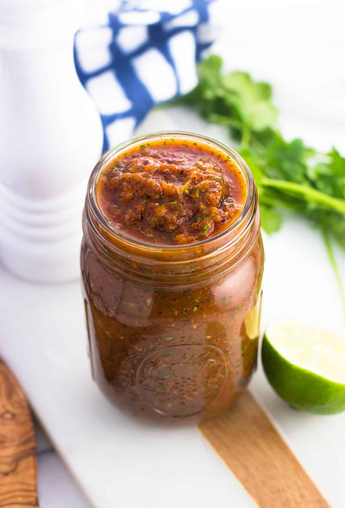 A glass mason jar filled with salsa on a marble board next to a lime half, a salt shaker, and a bunch of cilantro.