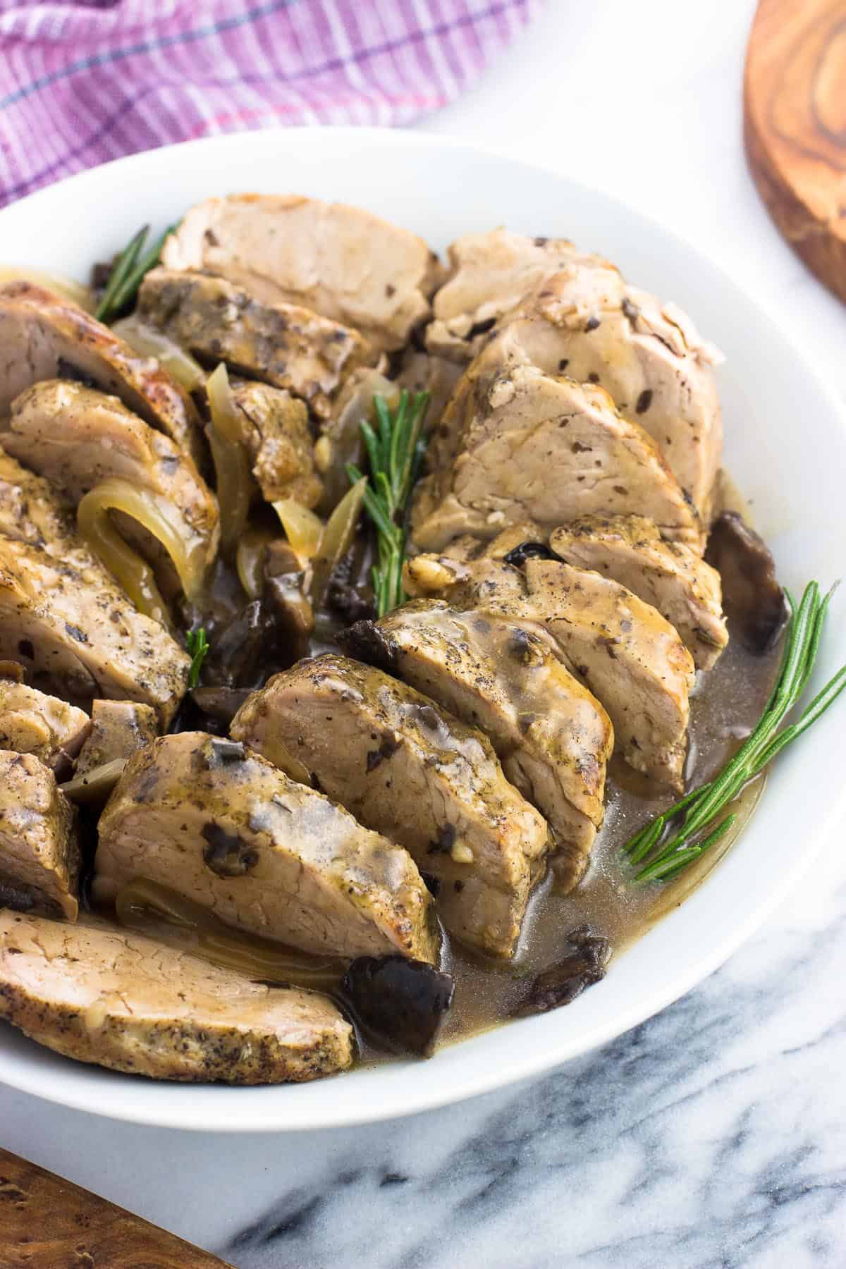 Sliced pork tenderloin in a serving dish garnished with sauce and fresh rosemary sprigs.