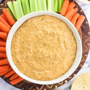 An overhead shot of buffalo dip in a ceramic serving bowl on a board surrounded by baby carrots and celery sticks