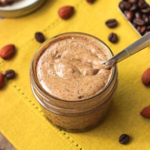 A small glass jar filled with almond butter on a placemat with scattered almonds and espresso beans