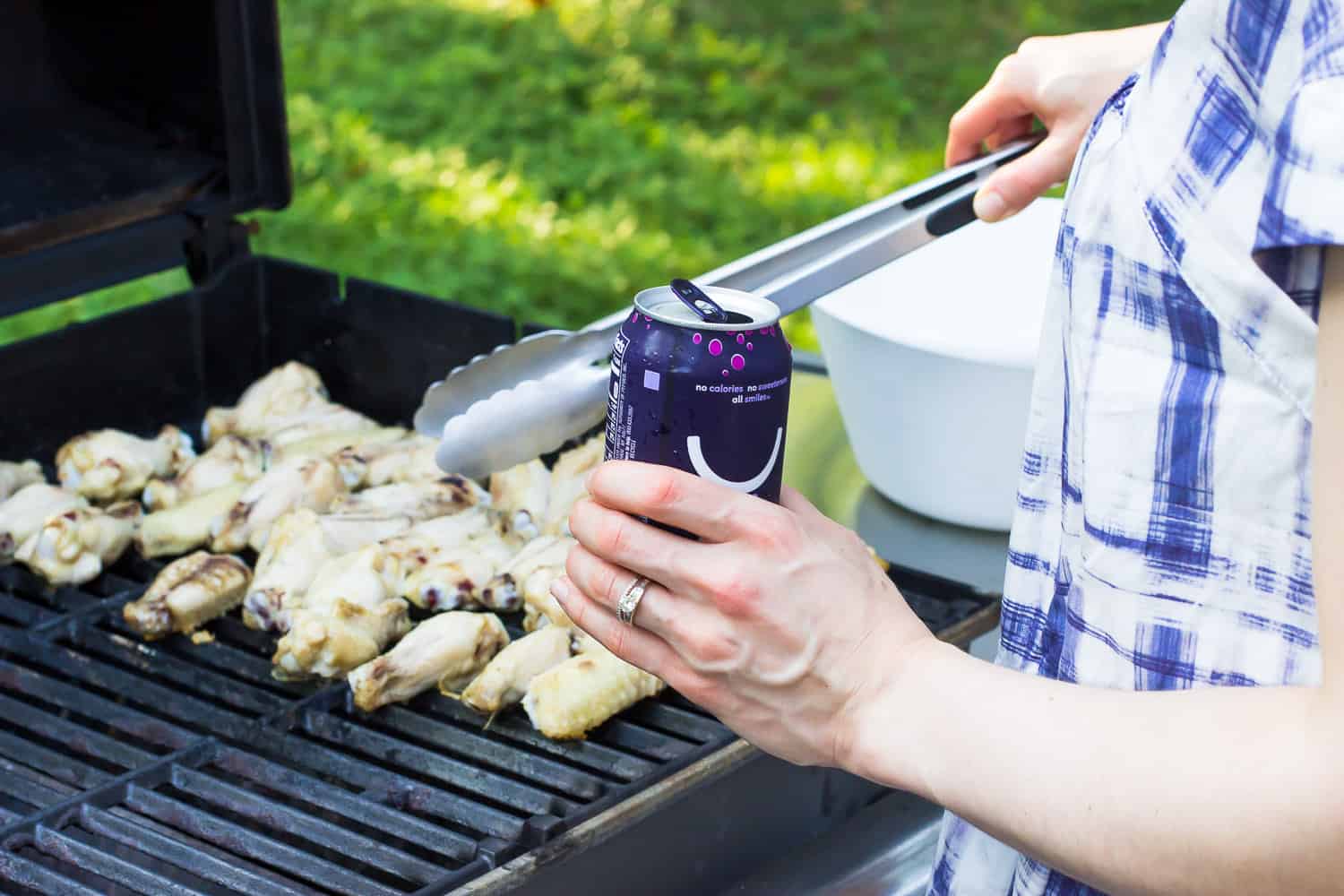 A woman cooking wings on a grill holding a can of water.